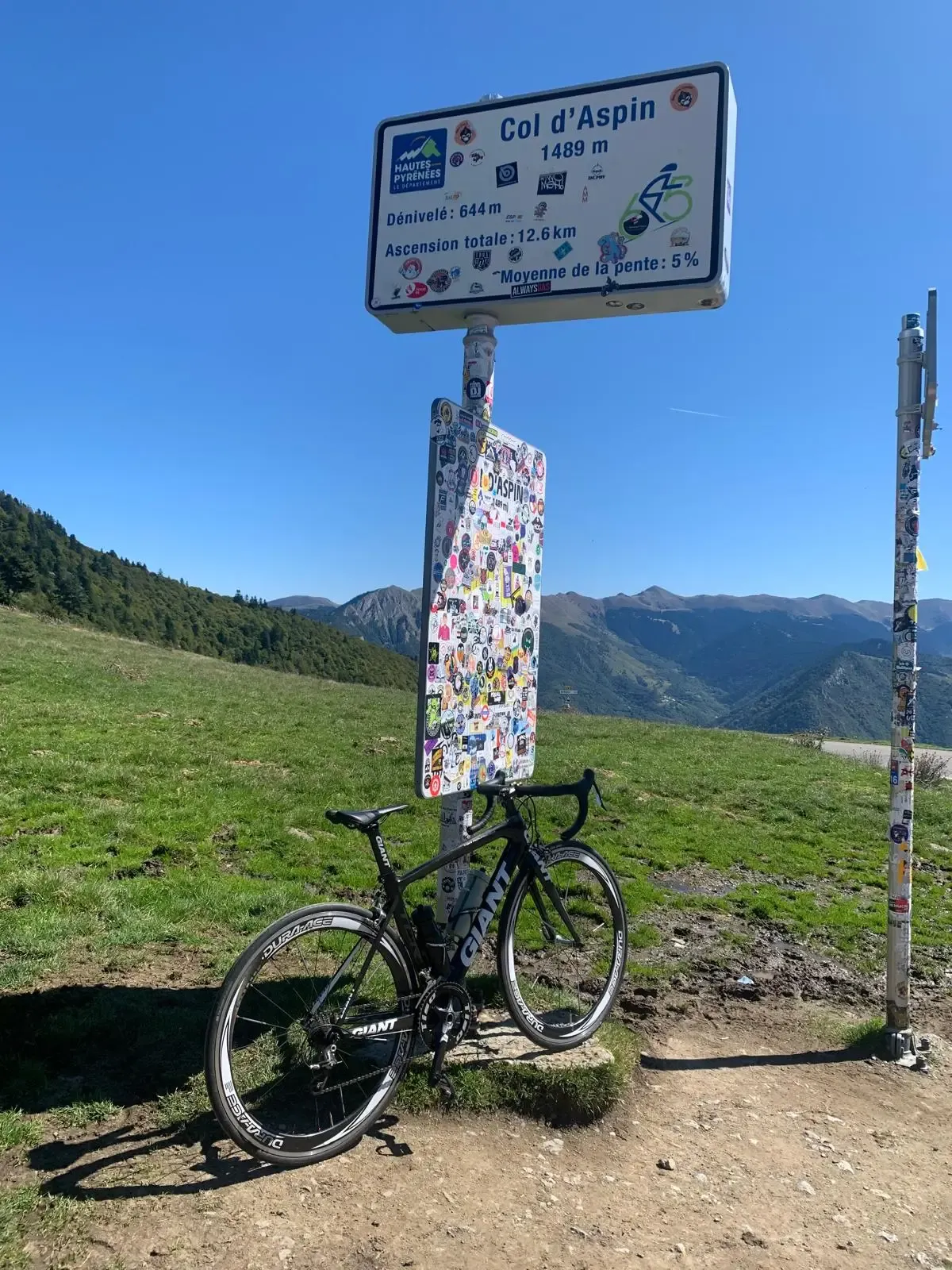 Vélo de route Giant noir appuyé contre le panneau officiel du Col d'Aspin (1489 m) au sommet. Le panneau est décoré de nombreux autocollants de clubs de cyclisme. En arrière-plan, un panorama ensoleillé sur les montagnes des Hautes-Pyrénées sous un ciel bleu clair